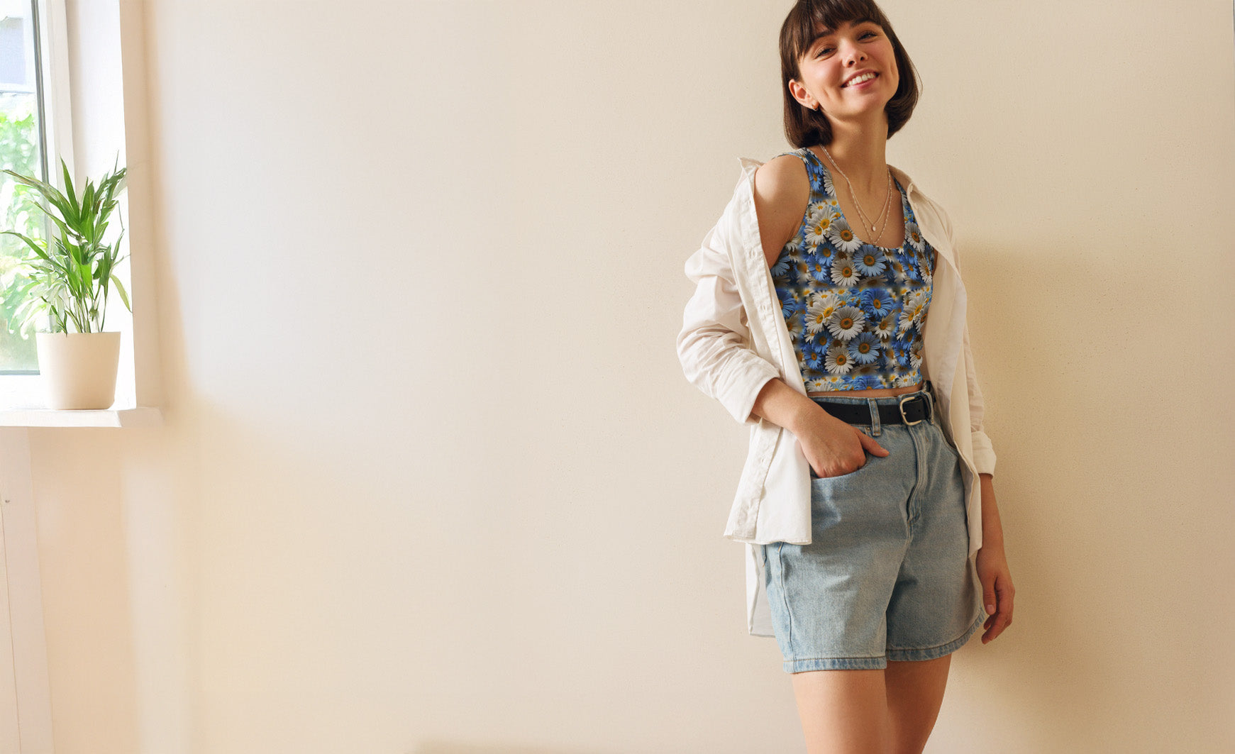 Woman wearing a floral top and denim shorts standing against a light-colored wall.