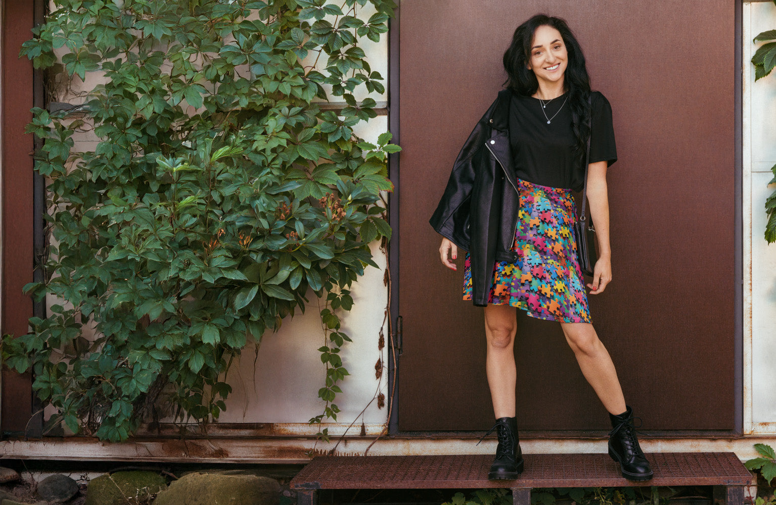 Woman standing in front of a brown wall with greenery, wearing a black jacket, black top, and colorful skirt.