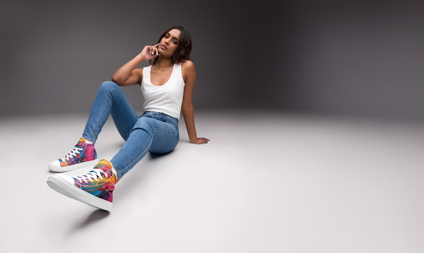 Woman sitting on a white surface wearing colorful sneakers, blue jeans, and a white tank top.